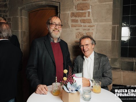 Zwei Männer stehen an einem runden Tisch vor einer Backsteinwand und lächeln in die Kamera. Auf dem Tisch befinden sich zwei Getränke mit gestreiften Strohhalmen und eine kleine Blumendekoration. Im Hintergrund sind weitere Gäste zu sehen. Auf der rechten Seite ist ein Fenster und das Logo von "United Kids Foundations" mit dem Hinweis "20 Jahre".