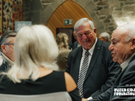 Eine Gruppe älterer Menschen steht in festlicher Kleidung in einem geschäftigen Raum. Ein Mann im Anzug lächelt freundlich, während er sich mit anderen unterhält. Im Hintergrund sind gemusterte Wanddekorationen und Steinwände zu sehen. Die Atmosphäre wirkt herzlich und gesellig. In der Ecke ist ein Logo mit dem Text „20 Jahre United Kids Foundations“ zu erkennen.