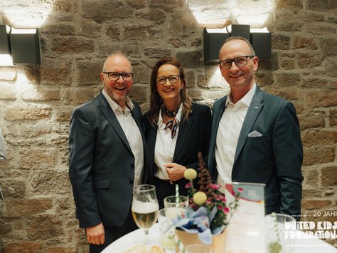 Drei Personen in formeller Kleidung stehen lächelnd vor einer Steinwand. Sie befinden sich in einer gemütlichen Atmosphäre, möglicherweise auf einer Veranstaltung. Vor ihnen steht ein Tisch mit mehreren Getränken und Blumenarrangement. Auf der unteren rechten Ecke ist ein Logo mit der Aufschrift „UNITED KIDS FOUNDATIONS – 20 JAHRE“.