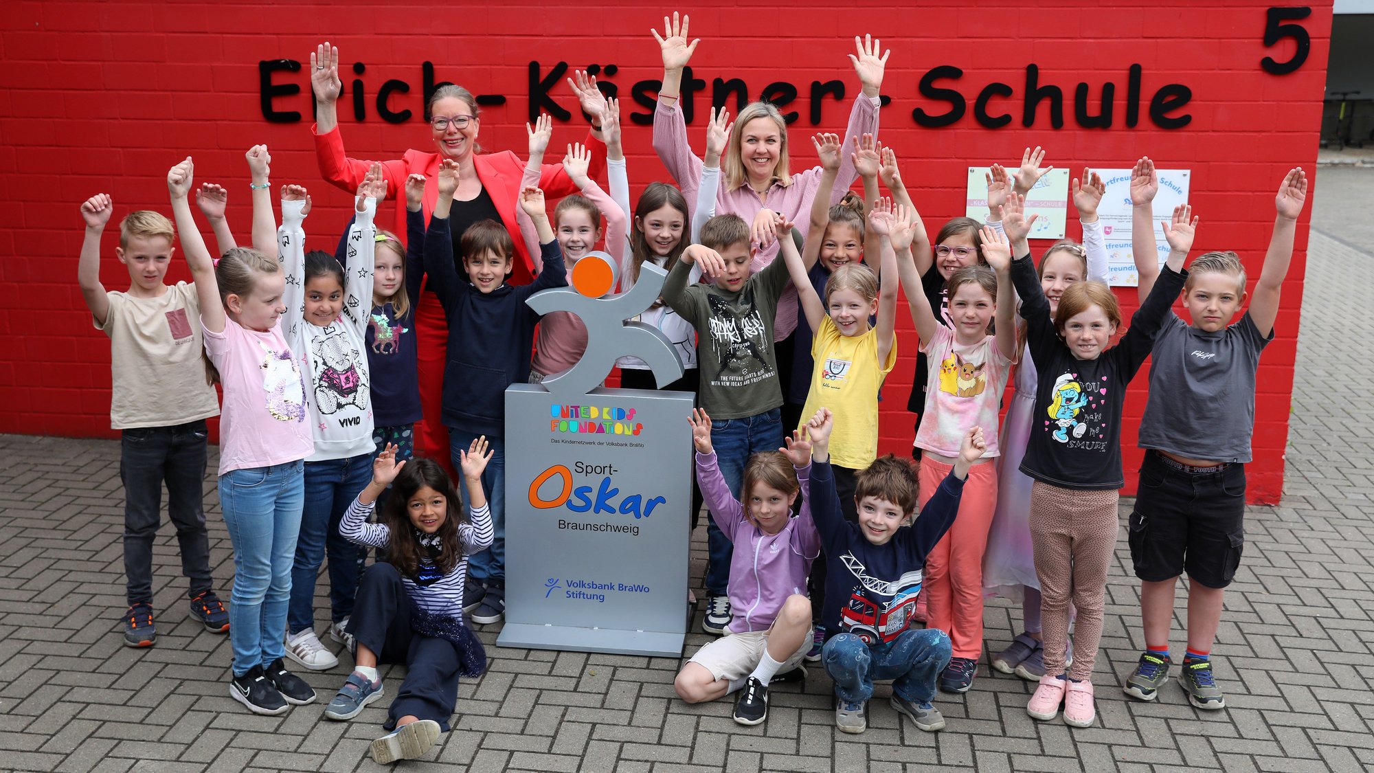 Eine Gruppe von Kindern und zwei Erwachsene stehen vor einer roten Wand mit der Aufschrift "Erich-Kästner-Schule" und strecken freudig die Arme in die Luft.