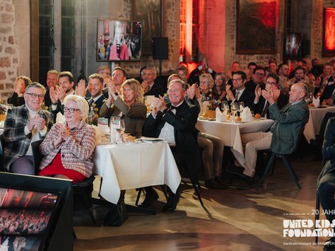 Eine Gruppe von gut gekleideten Menschen sitzt an runden Tischen in einem festlichen Saal. Sie klatschen und schauen nach vorne. Die Tische sind mit weißen Tischdecken, Weingläsern und Blumen dekoriert. Im Hintergrund sind alte Steinwände und Gemälde zu sehen. Ein Bildschirm zeigt eine Frau in rotem Outfit. Unten rechts im Bild steht "20 Jahre United Kids Foundations".