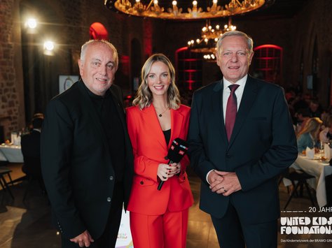 Drei Personen stehen in einem festlich beleuchteten Raum. Die zentrale Person trägt einen auffälligen roten Anzug und hält ein Mikrofon. Die beiden anderen Personen tragen dunkle Anzüge. Im Hintergrund sind gedeckte Tische mit Personen zu sehen. Eine Dekoration an der Wand zeigt den Schriftzug "20 Jahre United Kids Foundations".