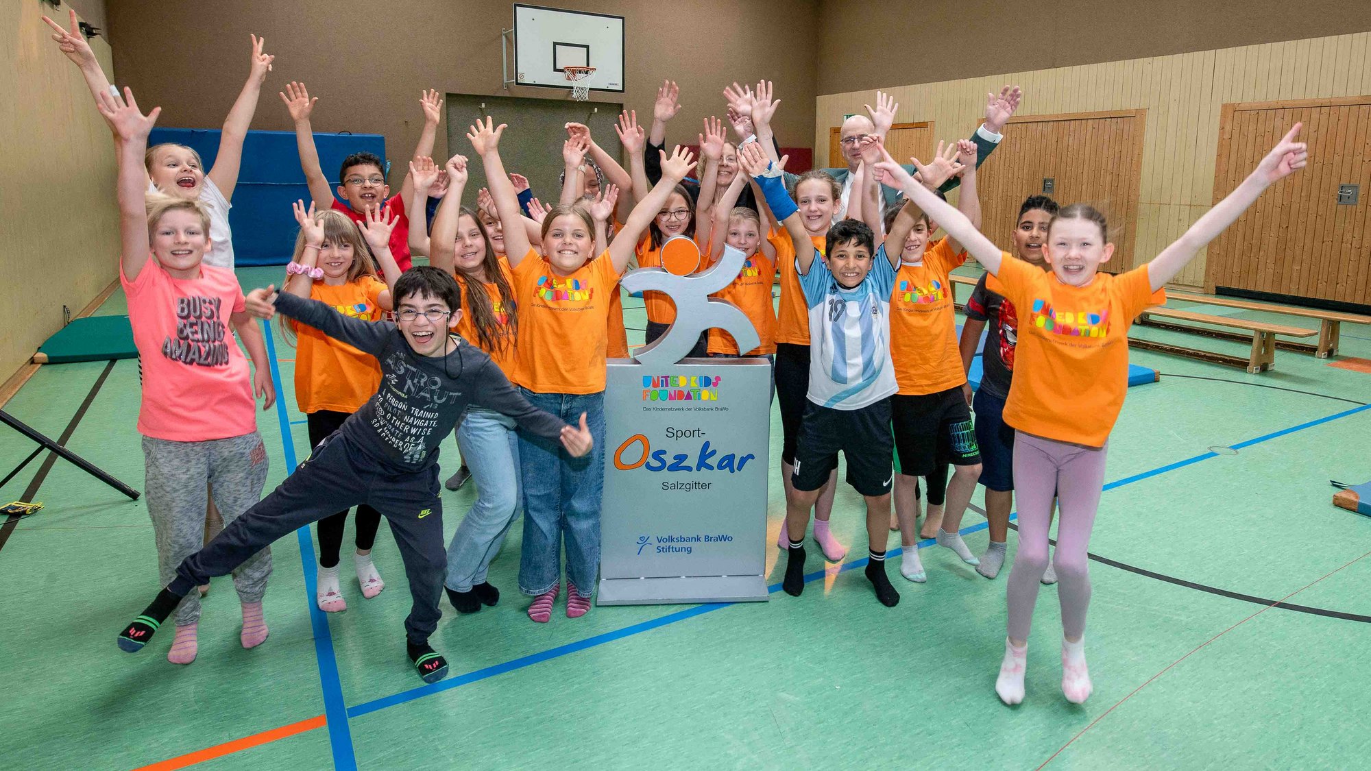 Eine Gruppe Kinder stehen freudig in einer Turnhalle und strecken ihre Arme in die Luft. In der Mitte steht die graue Sport-Oskar-Trophae.