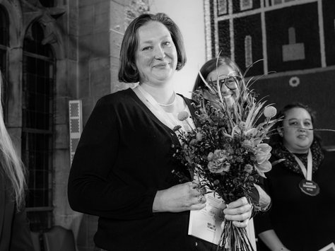 Eine Frau steht in einem Innenraum, vermutlich bei einer Veranstaltung. Sie hält einen Blumenstrauß in den Händen und lächelt. Neben ihr stehen zwei weitere Personen. Im Hintergrund sind ein großes Wandmosaik und ein gotisches Fenster zu sehen. In der unteren Ecke des Bildes ist das Logo der "United Kids Foundations" mit dem Schriftzug "20 Jahre" zu erkennen.
