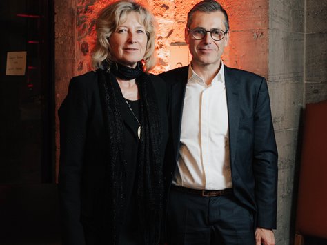 Eine Frau und ein Mann stehen nebeneinander in einem Raum mit einem Hintergrund aus Backstein und roter Beleuchtung. Die Frau trägt ein schwarzes Kleid mit einem schwarzen Schal, der Mann trägt einen dunklen Anzug mit einem weißen Hemd. Unten rechts im Bild ist das Logo von "United Kids Foundations" mit dem Schriftzug "20 Jahre".