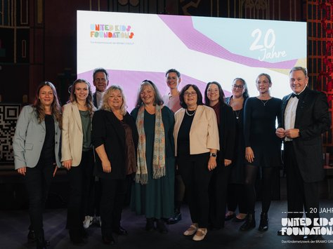 Eine Gruppe von Menschen steht zusammen auf einer Bühne vor einem großen Bildschirm. Der Bildschirm zeigt das Logo der "United Kids Foundations" und den Schriftzug "20 Jahre". Die Personen sind formell gekleidet. Die Veranstaltung scheint ein Jubiläum zu feiern, basierend auf der Dekoration und dem Schriftzug im Hintergrund.