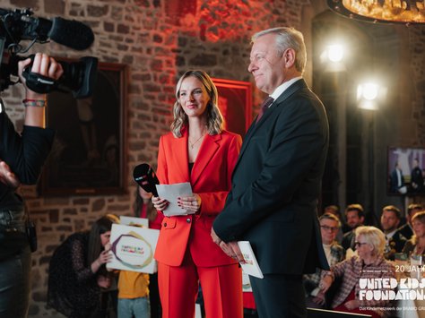 Zwei Personen, ein Mann und eine Frau, stehen nebeneinander in einem festlichen Raum mit Steinwänden. Die Frau trägt einen auffälligen roten Anzug und hält ein Mikrofon, während der Mann einen dunklen Anzug trägt. Beide scheinen auf einer Veranstaltung zu sprechen. Im Hintergrund sind unscharf Menschen zu sehen, die an Tischen sitzen sowie ein Kameramann, der die Szene filmt. Auf der rechten Seite ist ein TV-Bildschirm erkennbar. Rechts unten im Bild befindet sich ein Logo mit der Aufschrift "20 Jahre United Kids Foundations".