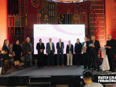 Eine Gruppe von Menschen steht auf einer Bühne vor einem großen Bildschirm. Der Hintergrund ist kunstvoll verziert und in warmem Licht beleuchtet. Ein Kameramann filmt die Szene, während einige Personen auf der Bühne Unterlagen halten und lesen. Am unteren rechten Rand des Bildes befindet sich das Logo der "United Kids Foundations" mit dem Hinweis auf ihr 20-jähriges Jubiläum.