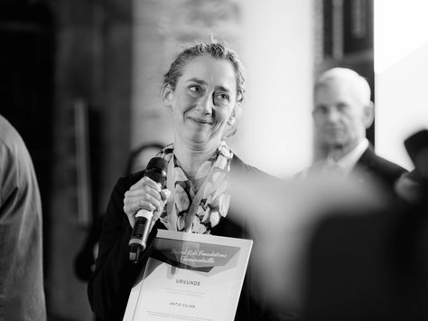 Eine Frau hält ein Mikrofon und eine Urkunde, während sie auf einer Bühne steht. Sie trägt einen Blazer und einen gemusterten Schal. Im Hintergrund stehen unscharf andere Personen. Der Text auf der Urkunde ist teilweise lesbar. Es handelt sich um eine Veranstaltung der United Kids Foundations, wie in der Ecke des Bildes zu sehen ist. Das Bild ist in Schwarz-Weiß gehalten.