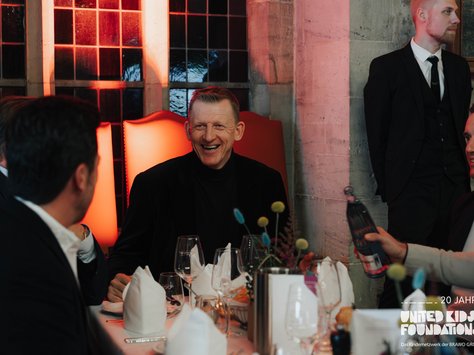 Ein eleganter Raum mit mehreren Personen an einem festlich gedeckten Tisch. Im Mittelpunkt sitzt ein Mann in dunkler Kleidung, der lächelnd mit den anderen am Tisch interagiert. Vor ihm stehen Weingläser und weiß gefaltete Servietten. Im Hintergrund stehen große Fenster und ein Mann im Anzug. Der Raum ist mit warmem Licht beleuchtet. Auf der rechten unteren Ecke ist das Logo "United Kids Foundations" zu sehen.