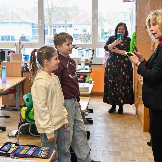 In einem Klassenzimmer steht eine Frau in einem schwarzen Anzug und hält eine grüne Figur in den Händen, während sie mit zwei stehenden Kindern spricht. Eine Frau im Hintergrund fotografiert die Szene mit einem Smartphone. Weitere Kinder sitzen an ihren Tischen, während auf dem Boden ein buntes Federmäppchen liegt. Im Raum herrscht eine freundliche, lernfreudige Atmosphäre.
