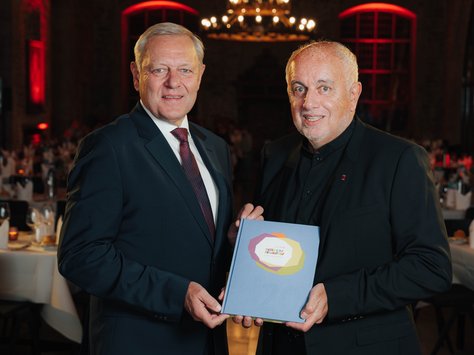 Zwei Männer in Anzügen stehen in einem festlich beleuchteten Raum mit gedeckten Tischen. Beide halten gemeinsam ein Buch, auf dem "United Kids Foundations" steht. Im Hintergrund sind große Fenster und ein Kronleuchter zu sehen. Es scheint sich um eine Veranstaltung zum 20-jährigen Jubiläum der United Kids Foundations zu handeln.