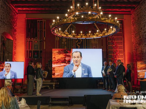 In einem prächtig beleuchteten Raum mit Steinwänden und dekorativen Wandteppichen steht ein großer Bildschirm, der das Bild eines Mannes zeigt, der in einem Anzug spricht. Eine Gruppe von Menschen steht auf der Bühne und hört zu. Ein massiver Kronleuchter hängt von der Decke. Im Vordergrund sitzen Personen an einem Tisch. Das Logo "United Kids Foundations" ist in der unteren rechten Ecke des Bildes zu sehen.