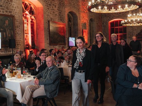 In einem festlich beleuchteten Raum mit steinernen Wänden und Holzbalken-Decke findet eine Veranstaltung statt. An runden Tischen sitzen Menschen, die aufmerksam einer Präsentation folgen und teilweise klatschen. Im Vordergrund geht eine Frau in heller Hose und Schal, gefolgt von anderen Personen, durch den Raum. An den Wänden hängen Gemälde und von der Decke hängen große Kronleuchter. Auf dem Bildrand ist ein Logo der "United Kids Foundations" mit dem Hinweis auf ein 20-jähriges Jubiläum zu sehen.