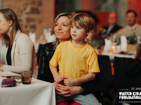 Eine Frau sitzt mit einem kleinen Jungen auf dem Schoß an einem Tisch, der mit einem weißen Tischtuch bedeckt ist. Der Junge trägt ein gelbes T-Shirt und die Frau hat einen sanften Ausdruck im Gesicht. Menschen im Hintergrund sitzen ebenfalls an Tischen und der Raum scheint festlich dekoriert zu sein. Auf dem Bild ist oben rechts ein Logo von United Kids Foundations mit dem Text "20 Jahre".