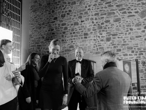 Eine Gruppe von Menschen steht in einem Raum mit einer Backsteinwand. Die Szene wirkt festlich, einige Personen lächeln und unterhalten sich. Eine Frau im Vordergrund scheint gerührt oder erfreut zu sein. Im Hintergrund ist eine helle Leuchttafel zu sehen, darauf die Aufschrift "20 Jahre United Kids Foundations", darunter steht "Das Kindernetzwerk der BRAWO GROUP". Atmosphärisches Schwarz-Weiß-Foto.