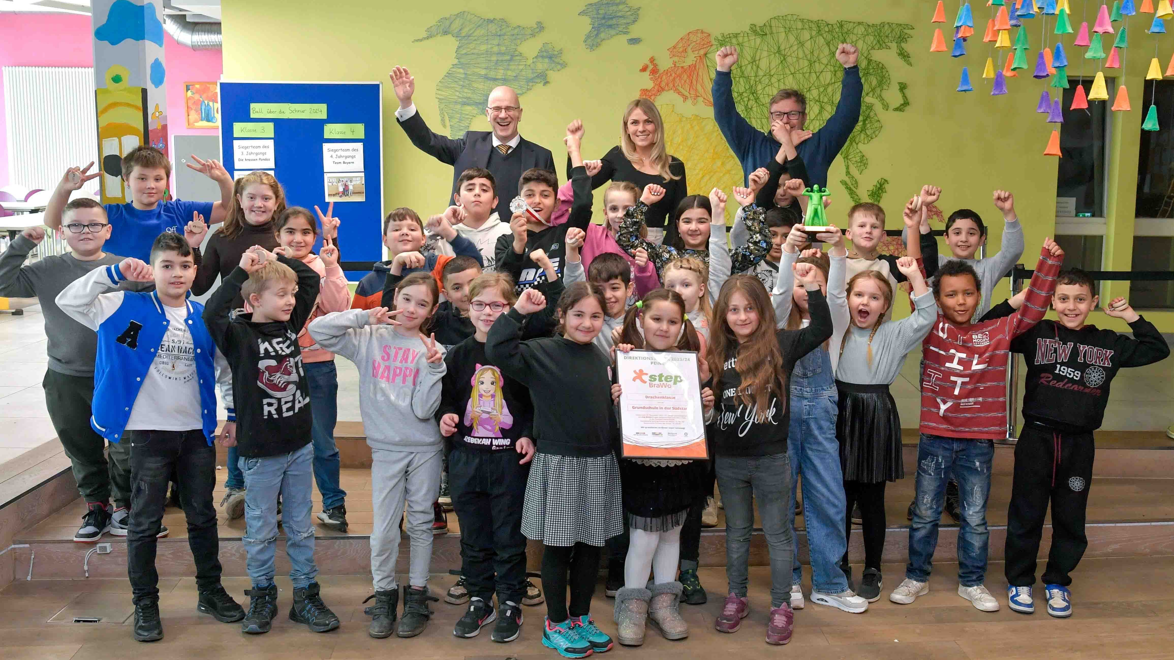 Eine Gruppe von Kindern und drei Erwachsene stehen lächelnd vor einer gelben Wand mit bunter Weltkarte. Sie strecken die Arme in die Luft.