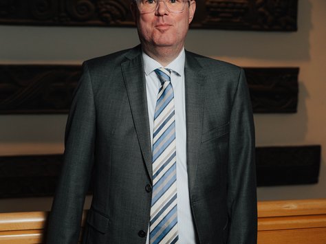 Ein Mann in einem grauen Anzug steht in einem Innenraum. Er hat eine Brille und trägt eine gestreifte Krawatte. Im Hintergrund sind dekorative Holzschnitzereien zu sehen, und über seinem Kopf fällt warmes Licht auf die Wand. In der unteren rechten Ecke des Bildes befindet sich das Logo von "United Kids Foundations" zum 20-jährigen Jubiläum.