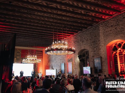 Eine große Veranstaltung in einem historischen Gebäude mit einer Steinwand und gotischen Fenstern, die in warmem Rot beleuchtet sind. Der Raum ist voll mit elegant gekleideten Gästen, die einem Auftritt auf der Bühne folgen. Zwei große, kreisförmige Kronleuchter hängen von der Decke, die mit Holzbalken verziert ist. Rechts im Bild sieht man einen Bildschirm mit dem Logo "United Kids Foundations".