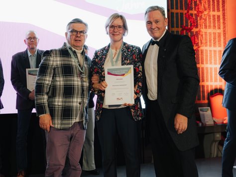 Auf dem Bild sind drei Personen vor einem Hintergrund mit dekorativem Muster und einer Projektion mit der Aufschrift "20 Jahre" zu sehen. Die Person in der Mitte hält eine Urkunde in den Händen. Alle drei tragen formelle Kleidung. Im Hintergrund steht eine weitere Person. Unten rechts im Bild ist das Logo von "United Kids Foundations" mit dem Hinweis auf das 20-jährige Jubiläum abgebildet.