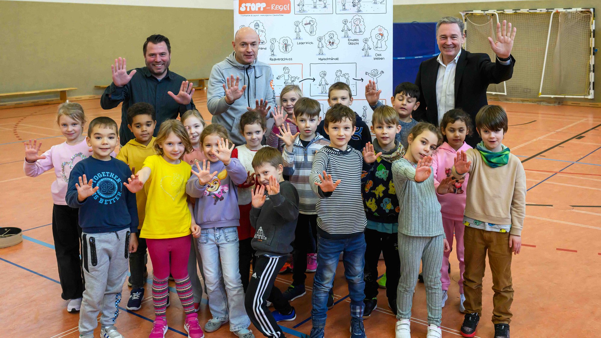 Das Bild zeigt eine Gruppe mit Kindern die in die Kamera winken. Sie stehen zusammen mit drei Erwachsenen in einer Turnhalle vor einem erklärenden Plakat.