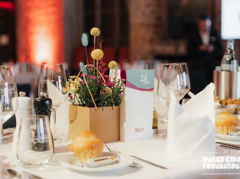 Ein elegant gedeckter Tisch in einem festlichen Raum. In der Mitte steht ein Blumengesteck in einem einfachen Topf, umgeben von Weingläsern, Besteck und gefalteten Stoffservietten. Auf dem Tisch liegt eine Karte mit der Aufschrift „20 Jahre“. Im Hintergrund ist eine Person unscharf zu erkennen. Der Raum ist in warmen Tönen beleuchtet und vermittelt eine einladende Atmosphäre.