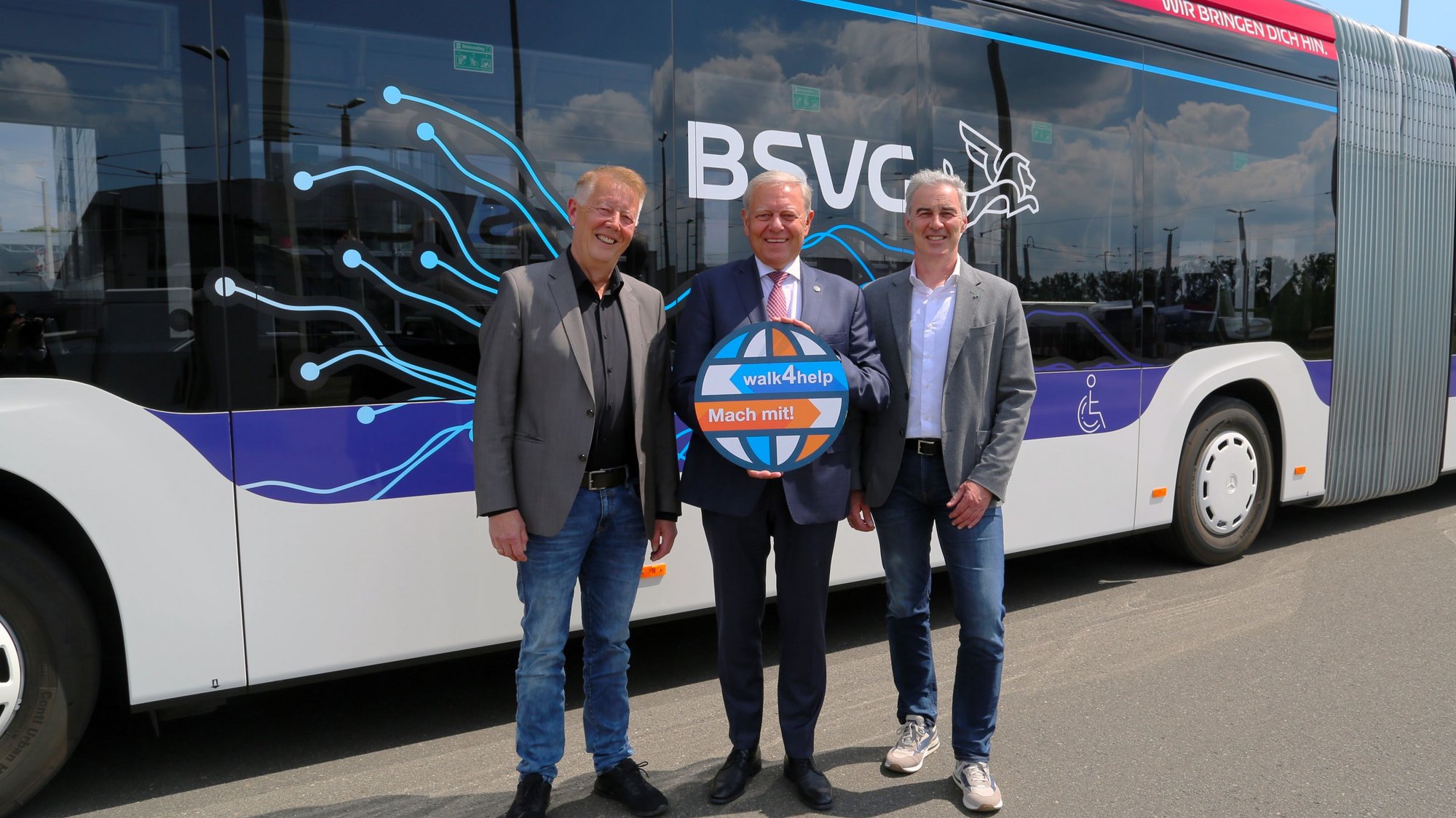 Jörg Reincke und Ralf Sygusch, Geschäftsführer des Verkehrsverbunds Region Braunschweig stehen links und rechts neben Jürgen Brinkmann, der das Logo des walk4help vor der Brust in der Hand hält. Sie stehen vor einem Bus der BSVG.