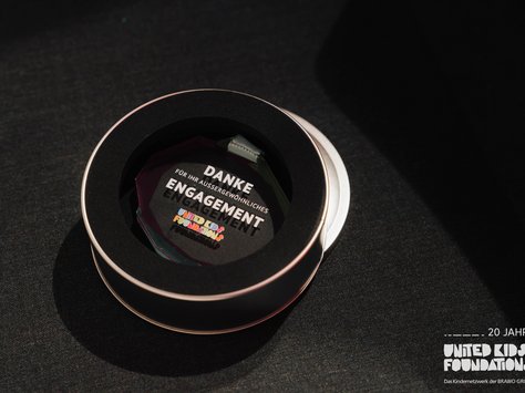 Ein rundes Etui mit einem innenliegenden, dekorativen Schild, das eine Danksagung für außergewöhnliches Engagement zeigt. Darauf ist auch das Logo der "United Kids Foundations" zu sehen. Das Bild feiert das 20-jährige Jubiläum der Organisation. Der Hintergrund ist dunkel gehalten, wodurch das Etui und das Schild besonders hervorgehoben werden.