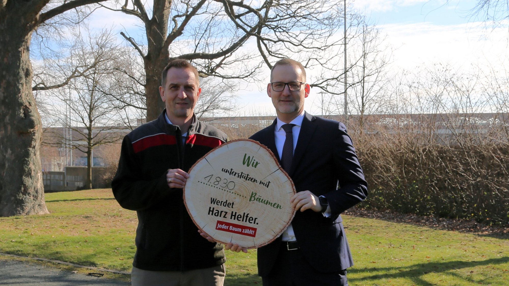 Zwei Männer stehen in einem Park. Sie halten gemeinsam eine runde Holzscheibe mit der Aufschrift „Wir unterstützen mit 1.830 Bäumen. Werdet Harz Helfer. Jeder Baum zählt!“. 