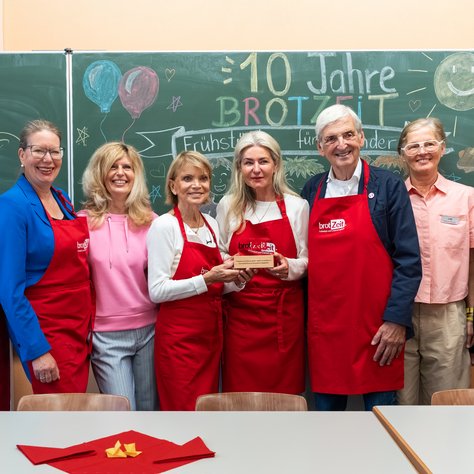 Eine Gruppe von acht Personen steht lächelnd vor einer Tafel und trägt rote Schürzen mit der Aufschrift "brotZeit". Zwei Frauen in der Mitte halten gemeinsam eine hölzerne Trophäe. Auf der Tafel im Hintergrund steht "10 Jahre BROTZEIT" umgeben von bunten Zeichnungen, darunter Luftballons und eine Sonne.