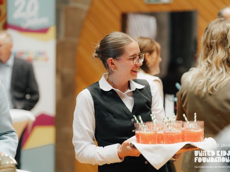 Eine Frau mit Brille und zusammengebundenem Haar trägt ein Tablett mit mehreren rosa Getränken, die mit gestreiften Strohhalmen versehen sind. Sie trägt ein weißes Hemd und eine schwarze Weste. Im Hintergrund sind unscharf stehende und sitzende Personen zu sehen, sowie ein Schild mit der Aufschrift "20 Jahre UNITED KIDS FOUNDATIONS". Die Stimmung wirkt fröhlich und lebendig.