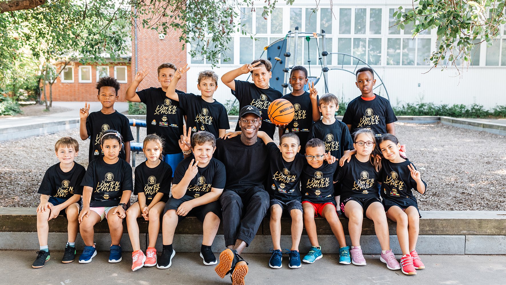 Auf dem Bild ist eine Gruppe Kinder in kurzen Hosen und schwarzen Shirts. Die Kinder in der ersten Reihe sitzen nebeneinander, in der Mitte sitzt ein Erwachsener Mann. Dahinter stehen ebenfalls Kinder in einer Reihe, von denen einer einen Basketball hochhält. Im Hintergrund ist ein Gebäude zu sehen.