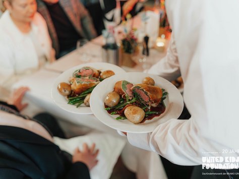 Eine Bedienung serviert zwei gefüllte Teller mit einer kunstvollen Anordnung von Fleischstücken auf Bohnen und kleinen Ofenkartoffeln. Die Gäste sitzen an einem elegant gedeckten Tisch, bereit zu speisen. Im Hintergrund sind verschwommene Details von Gästen und Tischdekoration zu erkennen. Unten rechts ist ein Logo mit der Aufschrift "20 Jahre United Kids Foundations" zu sehen.