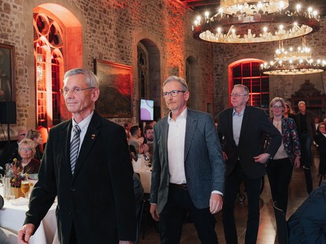 In einem festlich beleuchteten Raum mit Steinwänden und großen Kronleuchtern schreiten mehrere Personen in formeller Kleidung durch den Saal. Gäste sitzen an Tischen, die mit weißen Tischdecken und Gläsern gedeckt sind. Es herrscht eine feierliche Atmosphäre. Auf dem Boden ist das Logo der Veranstaltung "United Kids Foundations" sichtbar.