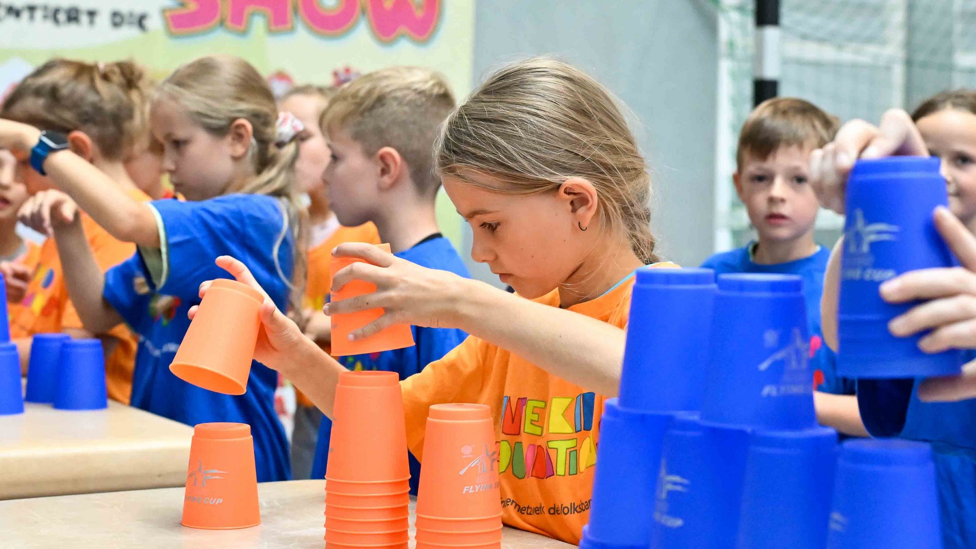 Ein großer Bewegungsparcours bildete den Mittelpunkt des Tages. Auch Geschicklichkeit und Reaktionsfähigkeit wurden dabei gefördert. Credit: Volksbank BRAWO/Isabell Massel In einer Reihe stehen Kinder in orangenen und blauen Tshirts vor Tischen, auf denen bunte Becher stehen, die sie konzentriert Türmen.