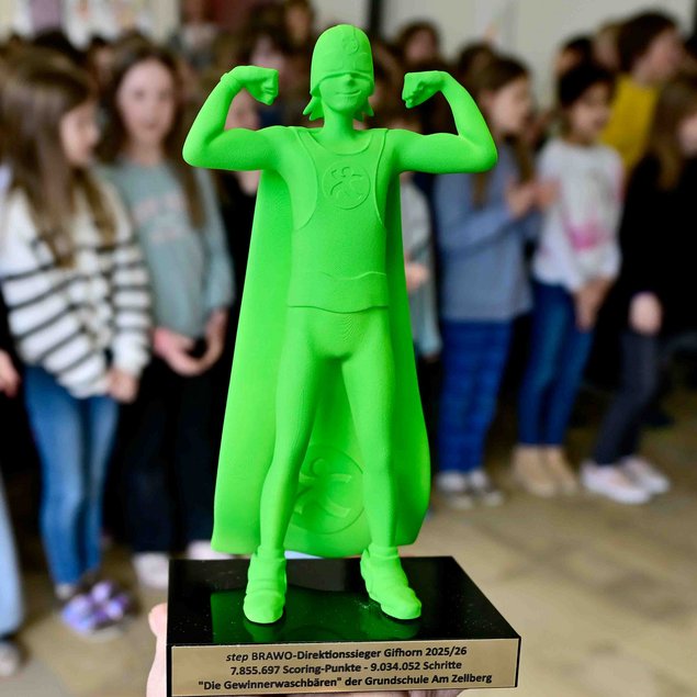 Das Bild zeigt eine grüne Trophäe in Form eines Superhelden mit Cape, der seine Muskeln anspannt. Im Hintergrund steht eine Gruppe von Kindern, die unscharf abgebildet sind. Auf dem Sockel der Trophäe ist eine Gravur zu sehen mit den Worten "step BRAWO-Direktonsessieger Gifhorn 2025/26" und weiteren Details.