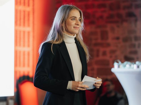 Eine Frau mit langen, blonden Haaren steht auf einer Bühne und hält Karten in der Hand. Sie trägt einen schwarzen Blazer und einen cremefarbenen Rollkragenpullover. Der Hintergrund zeigt eine Backsteinwand, die in warmem Rot beleuchtet ist. Rechts neben ihr steht ein hoher weißer Tisch mit einigen Deko-Elementen. In der unteren Ecke ist das Logo von "United Kids Foundations" und der Hinweis "20 Jahre" sichtbar.