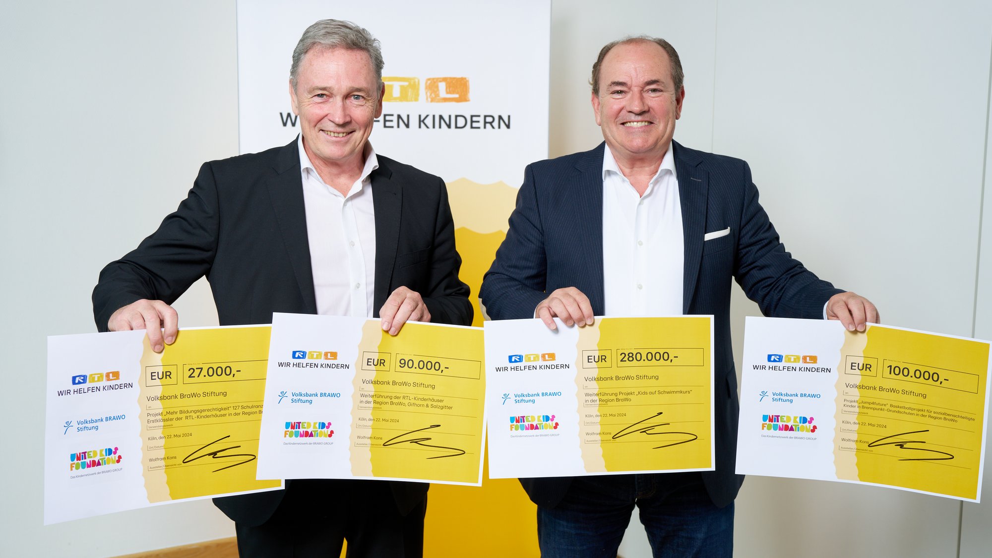 Thomas Fast (l.), Vorstandsvorsitzender der Volksbank BRAWO Stiftung, nahm die symbolischen Spendenschecks von Wolfram Kons, Vorstandsmitglied der "Stiftung RTL - Wir helfen Kindern e. V.", i. H. v. 470.000 Euro für Projekte in der BRAWO-Region entgegen. Zwei Männer im Anzug stehen lächelnd nebeneinander und halten in jeweils ihren Händen einen kleinen Spendencheck aus Papier.