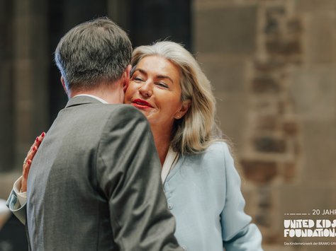 Eine Frau in einem hellblauen Outfit umarmt einen Mann in einem grauen Anzug. Die beiden scheinen sich freundlich zu begrüßen. Im Hintergrund ist ein unscharfer, dunkler Innenraum sichtbar. In der unteren rechten Ecke befindet sich das Logo von "United Kids Foundations" mit dem Hinweis auf das 20-jährige Jubiläum.