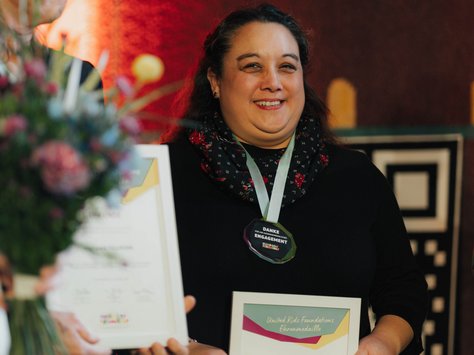 Eine Person hält eine Urkunde in der Hand und trägt eine Medaille um den Hals. Neben ihr steht eine weitere Person, die einen Blumenstrauß hält. Im Hintergrund ist eine bunte Wanddekoration zu sehen. Unten rechts befindet sich der Schriftzug "20 Jahre United Kids Foundations".