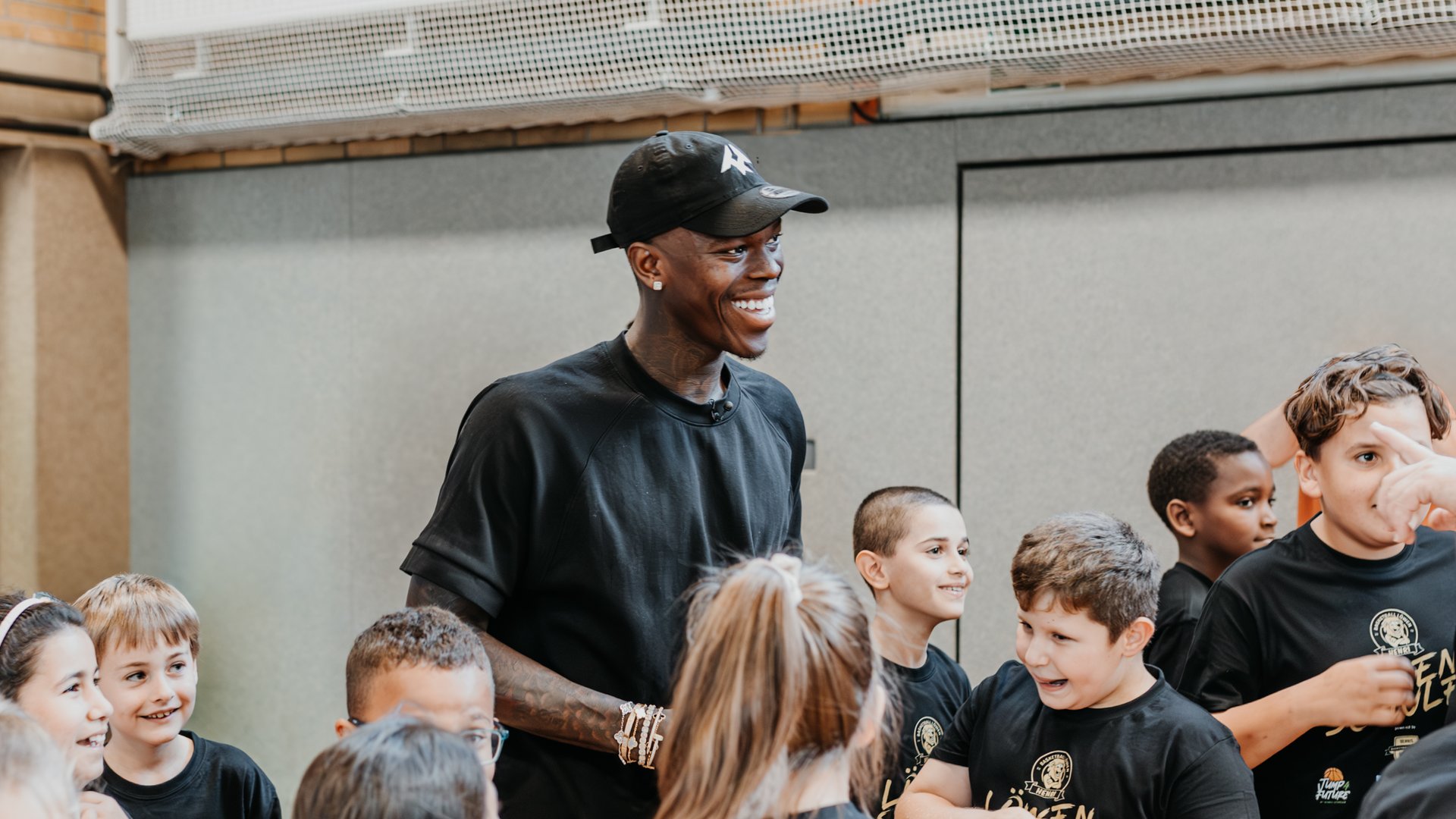 Eine Gruppe von Kindern in schwarzen T-Shirts mit der Aufschrift "Löwenschule" steht zusammen mit Dennis Schröder, der ein schwarzes T-Shirt und eine schwarze Kappe trägt. Schröder und die Kinder lachen und wirken fröhlich. Sie befinden sich in einer Halle mit hellem Boden und grauen Wänden.