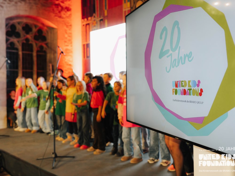 Eine Gruppe von Kindern steht auf einer Bühne und singt. Im Vordergrund ist ein Bildschirm zu sehen, auf dem das Logo der "United Kids Foundations" und der Text "20 Jahre" dargestellt sind. Der Raum hat historische Steinwände und große Fenster, die in warmem Licht erstrahlen.