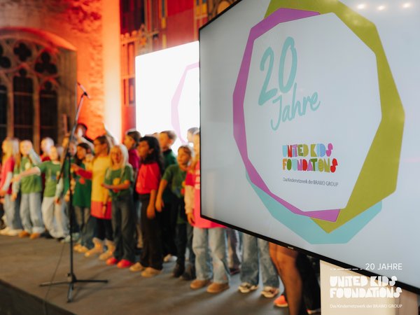 Eine Gruppe von Kindern steht auf einer Bühne und singt. Im Vordergrund ist ein Bildschirm zu sehen, auf dem das Logo der "United Kids Foundations" und der Text "20 Jahre" dargestellt sind. Der Raum hat historische Steinwände und große Fenster, die in warmem Licht erstrahlen.