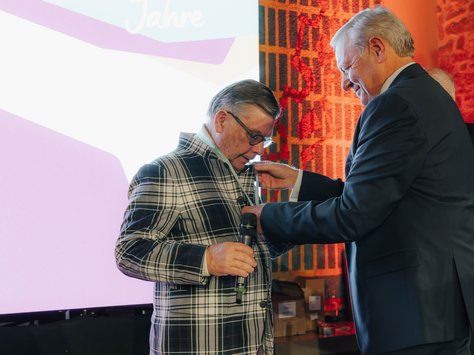 Zwei Männer stehen nebeneinander auf einer Bühne. Der eine im karierten Jackett hält ein Mikrofon, während der andere ihm eine Auszeichnung ans Revers heftet. Im Hintergrund ist ein großer Bildschirm mit der Aufschrift "20 Jahre" zu sehen. Die Atmosphäre wirkt festlich und feierlich.