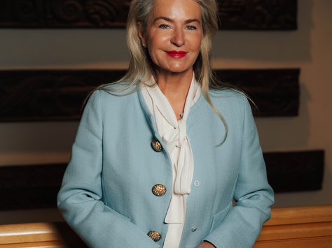 Eine Frau mit langen grauen Haaren steht lächelnd vor einem Holzhintergrund. Sie trägt einen hellblauen Blazer mit goldenen Knöpfen und eine weiße Bluse mit Schleife. Unten rechts im Bild befindet sich ein Logo der "United Kids Foundations", das ihr 20-jähriges Jubiläum verkündet.