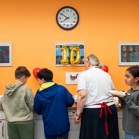 In einem Raum mit orangefarbener Wand stehen vier Personen vor einer Theke mit Snacks. Zwei Jungen schauen auf das Essen, während eine ältere Frau in einem weißen T-Shirt daneben steht. Ein Mädchen hält einen Teller und blickt zur Seite. Über der Theke hängt eine goldene Ballon-Zahl "10" unter einer Uhr. An der Wand sind mehrere gerahmte Bilder angebracht.