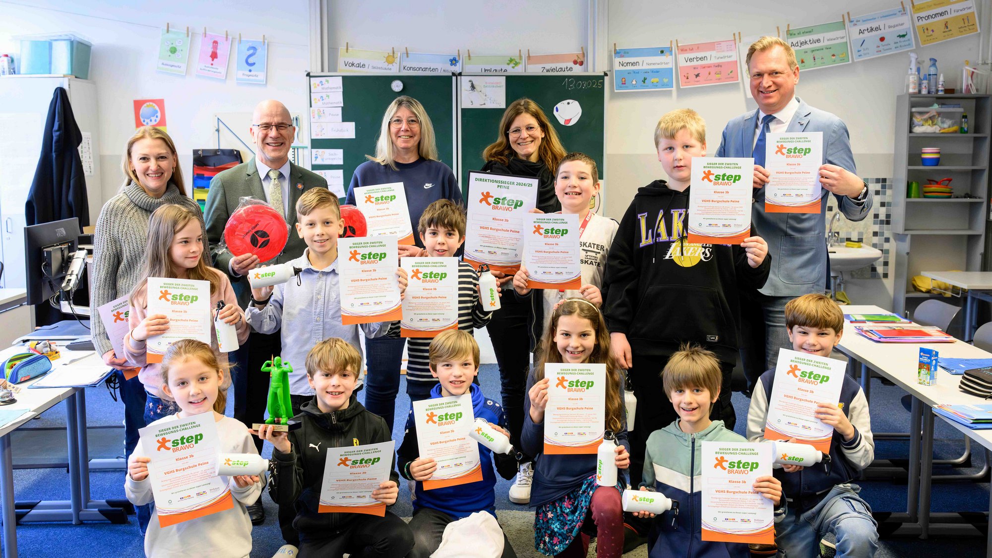 Eine Gruppe von Kindern und Erwachsenen steht in einem Klassenzimmer und hält stolz Urkunden in die Kamera. Einige Kinder halten zusätzlich kleine Preise wie eine grüne Superhelden-Trophäe oder einen roten Softball. Die Kinder sind fröhlich und feiern ihre Auszeichnung im Rahmen des „step BRAWO“-Programms. Im Hintergrund sind eine Tafel mit Unterrichtsmaterialien und eine Wäscheleine mit bunten Lernkarten zu sehen.