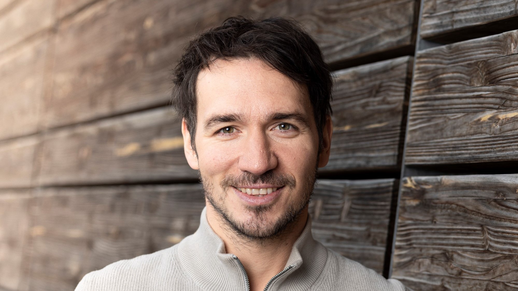 Ein Mann mit dunklen Haaren lächelt in die Kamera und lehnt sich entspannt an eine hölzerne, verwitterte Wand. Er trägt einen hellen, zippbaren Pullover. Die Szene wirkt warm und freundlich.