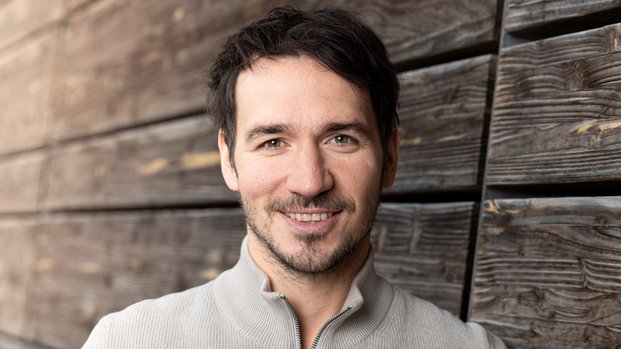 Ein Mann mit dunklen Haaren lächelt in die Kamera und lehnt sich entspannt an eine hölzerne, verwitterte Wand. Er trägt einen hellen, zippbaren Pullover. Die Szene wirkt warm und freundlich.