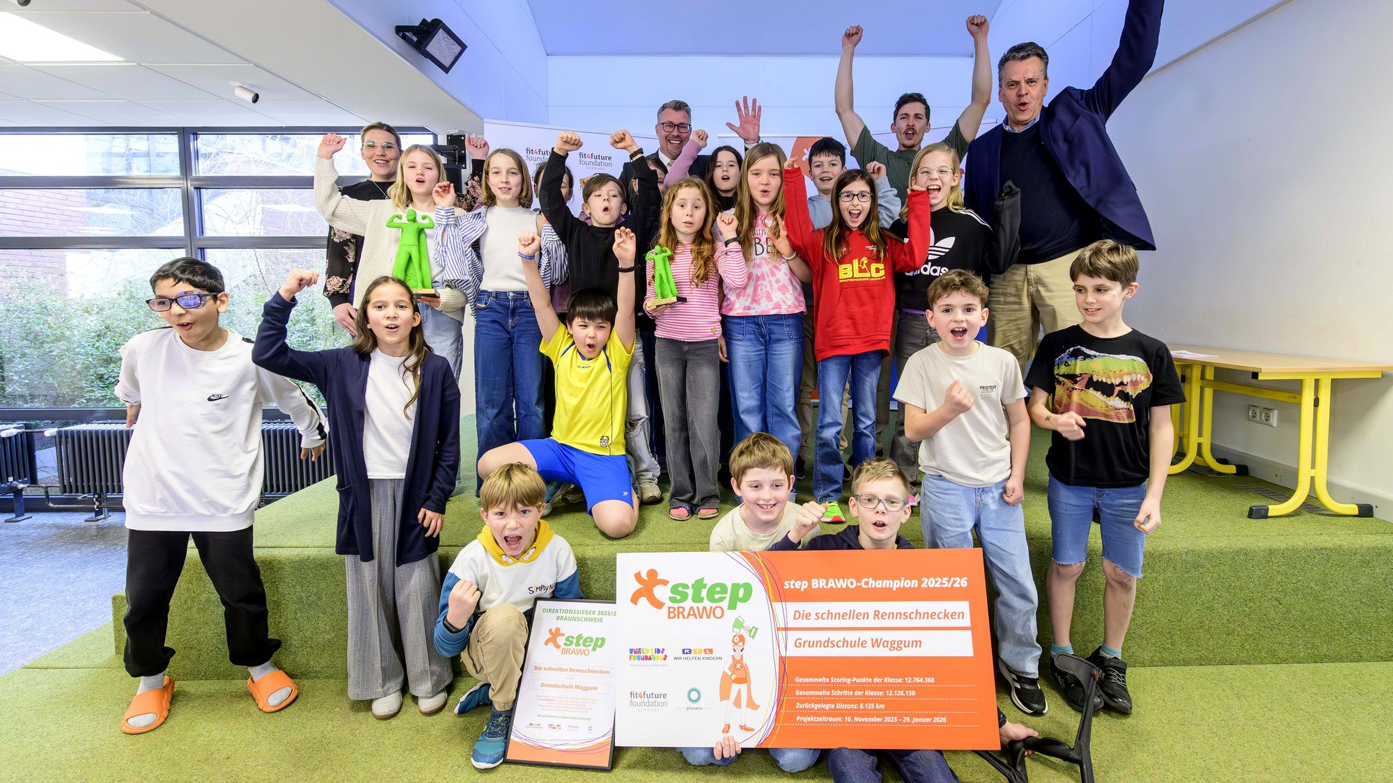 Eine Gruppe fröhlicher Kinder posiert zusammen mit einigen Erwachsenen in einem Raum auf einem Podest. Die Kinder tragen bunte Alltagskleidung und halten stolz ein großes orangenes Banner und eine Urkunde mit der Aufschrift "step BRAWO-Champion 2025/26". Einige Kinder haben ihre Fäuste in die Luft gereckt, offensichtlich in Feierstimmung.